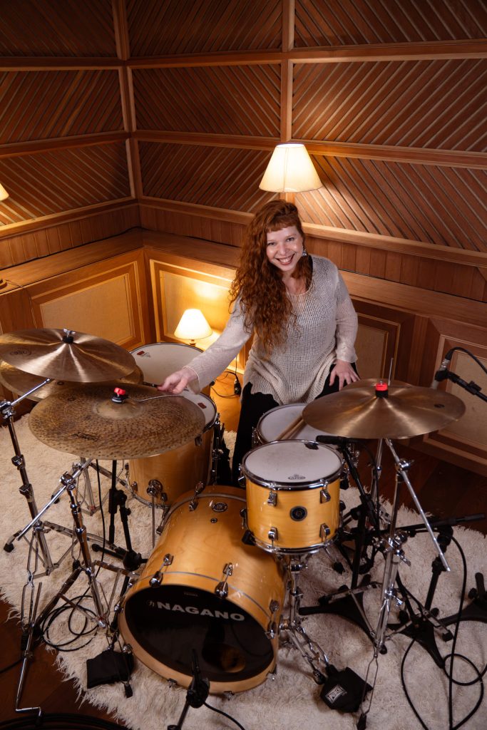 Caroline Calê, ruiva de cabelo cacheado, sentada diante de uma bateria.