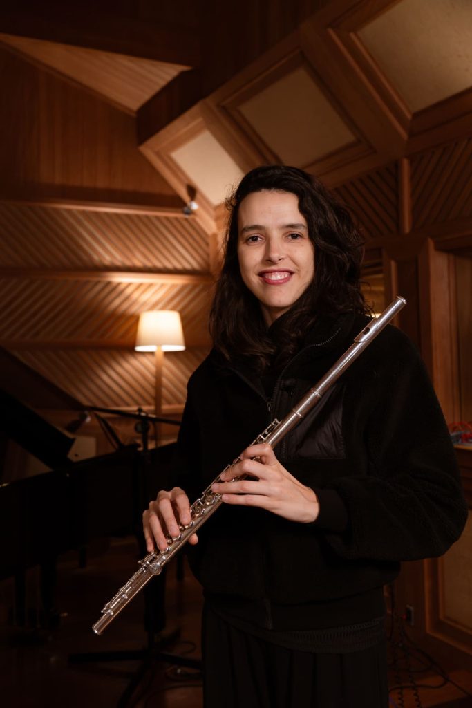 Larissa Galvão branca de cabelo escuro, segura uma flauta prateada.