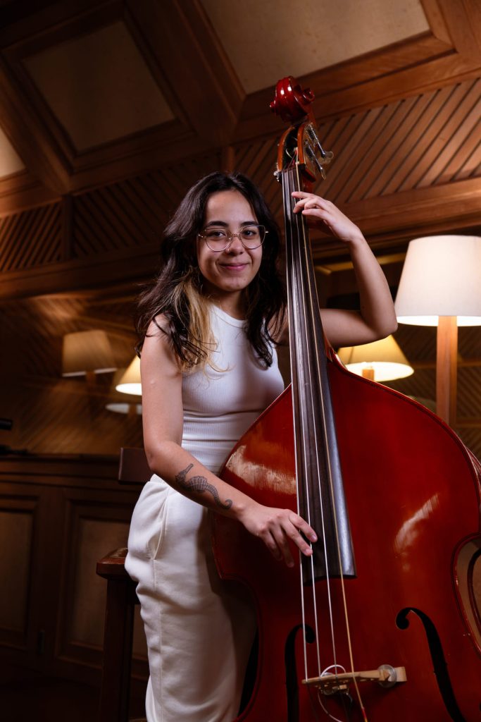 Laryssa Alves, branca de cabelo castanho liso, usa óculos e roupa branca, toca violoncelo.