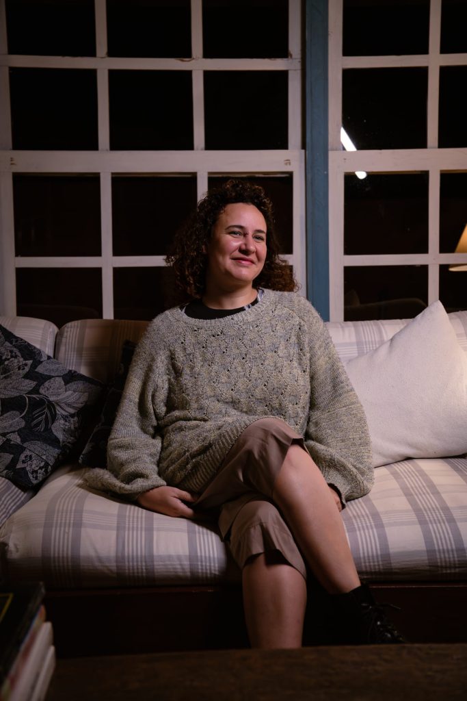 Marina Bastos, branca de cabelo cacheado, sentada em um sofá. Atrás, janelas brancas.