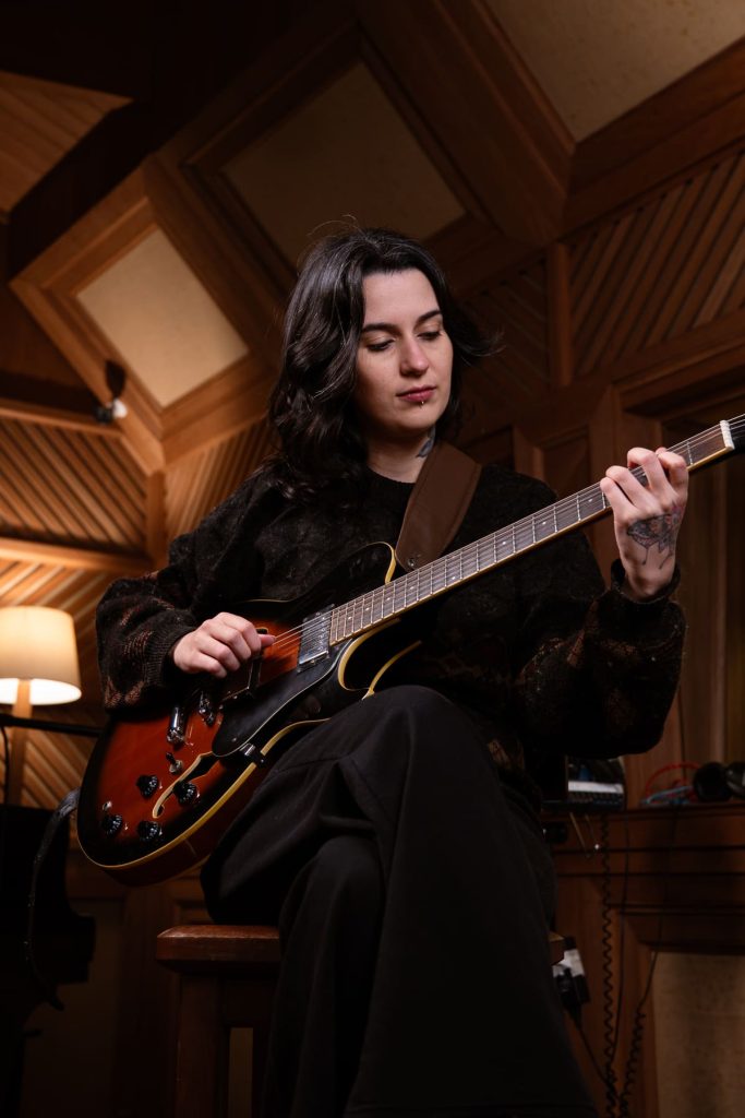 Miriam Momesso, branca de cabelo ondulado, toca guitarra.