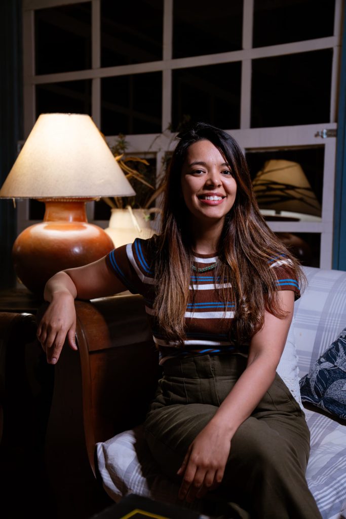 Monica Morais, parda de cabelo castanho liso, sentada em um sofá ao lado de um abajur aceso.