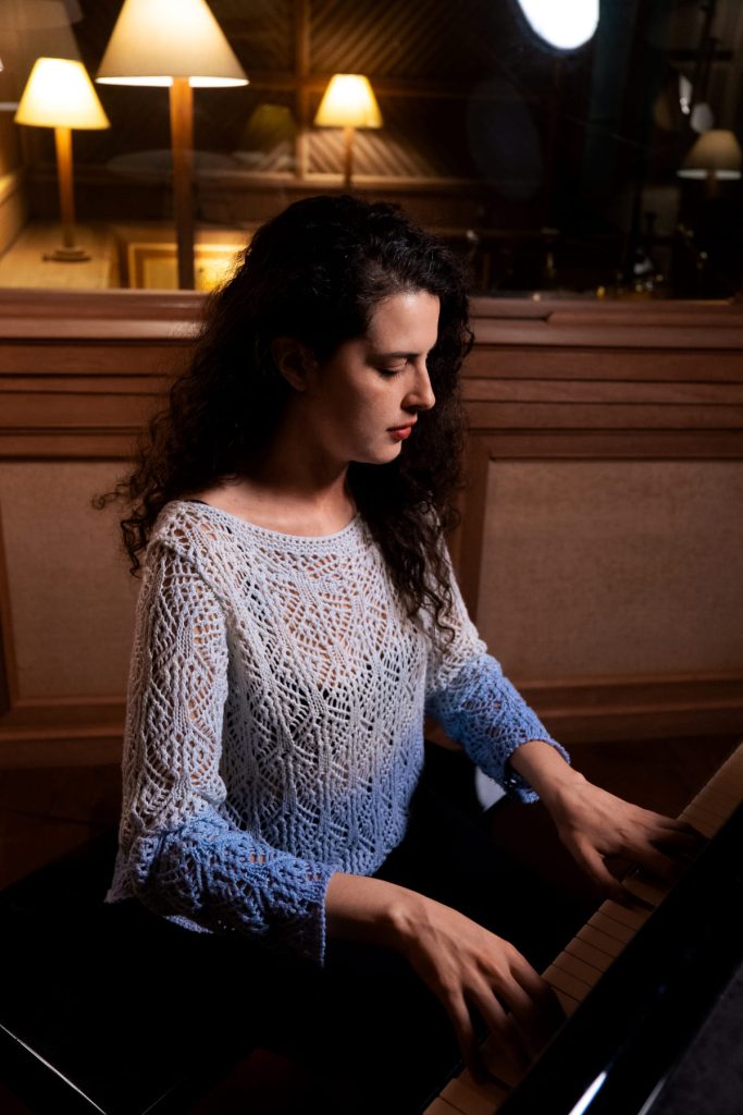 Viviane Pinheiro: branca de cabelo cacheado, com blusa de tricô branca e azul, toca piano.