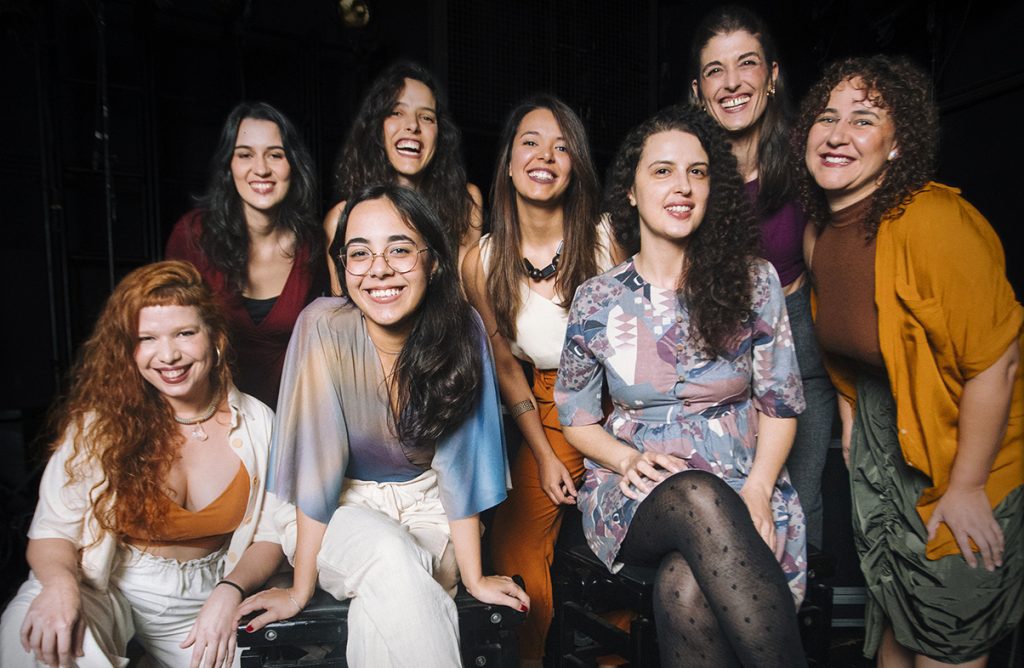 Foto institucional do grupo musical Octeta. Oito mulheres posam juntas em um palco escuro, algumas sentadas sobre um equipamento e outras em pé atrás. Todas sorriem para a câmera. O grupo veste roupas em tons terrosos, claros e estampados, em um ambiente de apresentação musical.