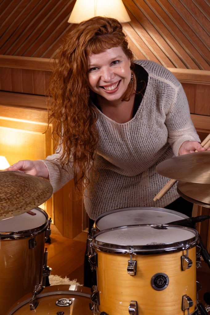 Caroline Calê, ruiva de cabelo cacheado, em pé diante de uma bateria. Ela veste tricô cinza claro e está com o tronco curvado para frente, segurando os pratos da bateria.