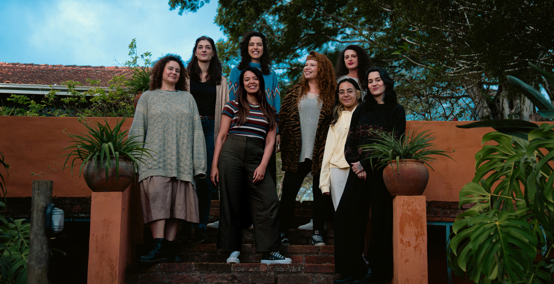 Foto institucional do grupo musical Octeta. Oito mulheres posam juntas em uma escadaria de tijolos, em ambiente externo com plantas e árvores ao fundo. As integrantes estão dispostas em diferentes alturas da escada e olham para a câmera, sorridentes.