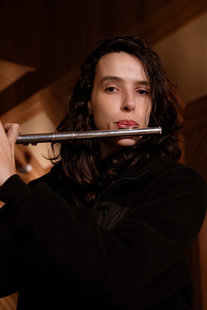 Larissa Galvão: branca de cabelo ondulado castanho,. Veste blusa preta e toca flauta.