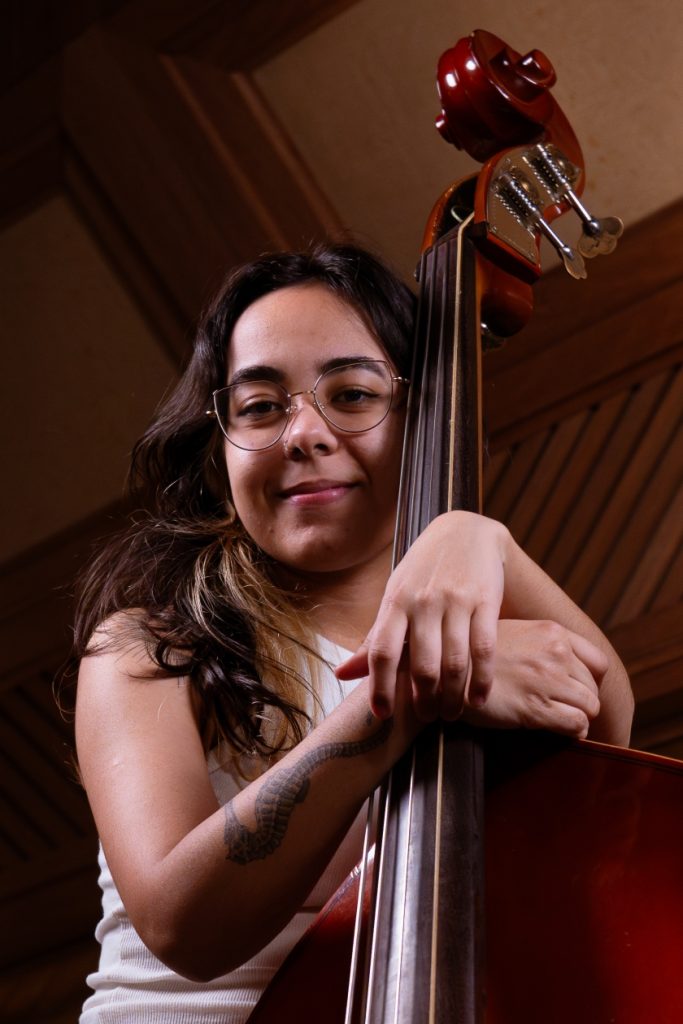 Laryssa Alves: branca de cabelo castanho liso, usa óculos e veste regata branca. Apoia as mãos em um violoncelo.