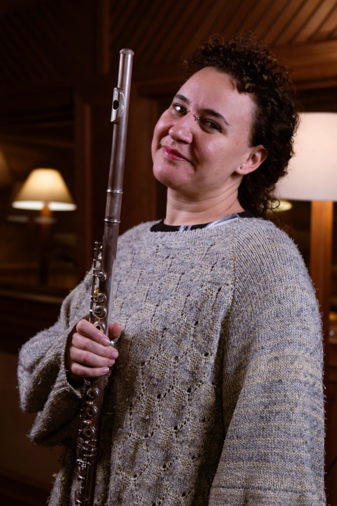 Marina Bastos: branca de cabelo cacheado, com piercing entre as sobrancelhas. Veste tricô cinza e segura uma flauta.