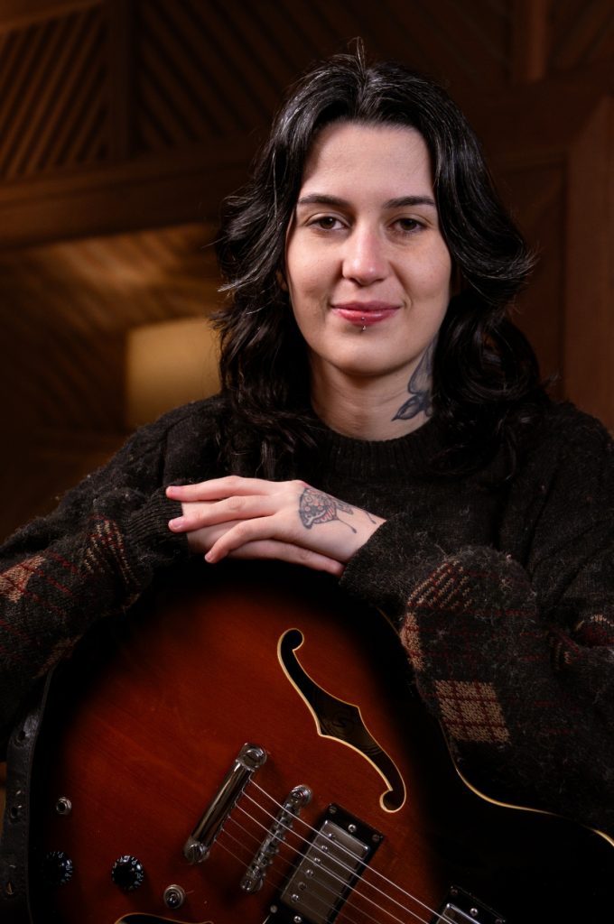 Miriam Momesso: branca de cabelo ondulado curto e com tatuagens no pescoço e dorso da mão. Veste tricô marrom e apoia as mãos em uma guitarra.