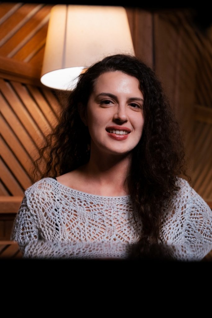 Foto: Viviane Pinheiro, branca de cabelo cacheado castanho longo. Veste blusa de crochê cinza.