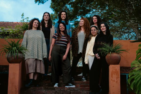 Foto institucional do grupo musical Octeta. Oito mulheres posam juntas em uma escadaria de tijolos, em ambiente externo com plantas e árvores ao fundo. As integrantes estão dispostas em diferentes alturas da escada e olham para a câmera, sorridentes.