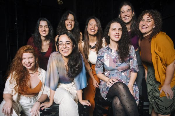 Foto institucional do grupo musical Octeta. Oito mulheres posam juntas em um palco escuro, algumas sentadas sobre um equipamento e outras em pé atrás. Todas sorriem para a câmera. O grupo veste roupas em tons terrosos, claros e estampados, em um ambiente de apresentação musical.